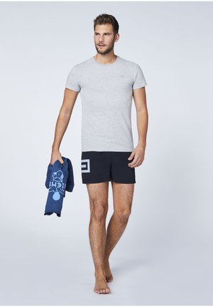 Gray short-sleeve t-shirt, navy swim shorts with graphic design, held navy towel. Model stands barefoot against a light background.