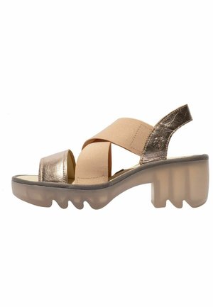 Sandalia abierta beige y metálica con tiras de tela cruzadas, suela translúcida gruesa y tacón ancho.