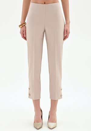 Donna che indossa pantaloni beige corti con dettagli a bottoni vicino all'orlo, braccialetti dorati e tacchi a punta dorati, in piedi su uno sfondo bianco.