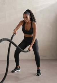 Svart sports-bh og leggings med teksturert overflate, kombinert med svarte joggesko. Personen bruker battle ropes mot en nøytral bakgrunn.