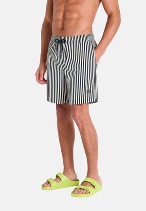 Uomo senza maglietta che indossa pantaloncini a righe verticali blu navy e bianchi con coulisse e sandali con fibbia verde neon, in piedi con le mani rilassate.