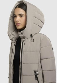 Lichtgrijze pufferjack met een grote capuchon, voorzien van doorgestikte kanalen, ritszakken en verstelbare toggles op de capuchon.
