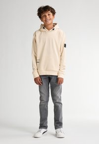 Sudadera de color beige con capucha y cordón ajustable, gran bolsillo frontal y parche negro como acento. Combinada con jeans grises y zapatillas blancas.