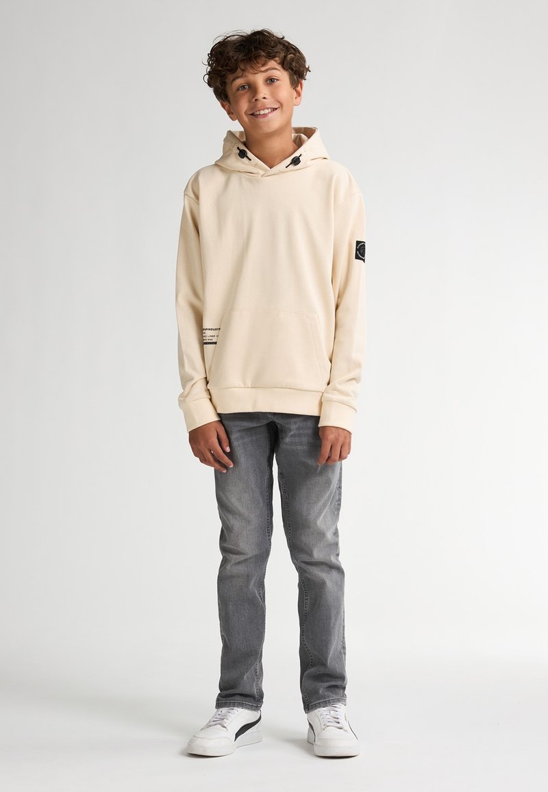Sudadera de color beige con capucha y cordón ajustable, gran bolsillo frontal y parche negro como acento. Combinada con jeans grises y zapatillas blancas.