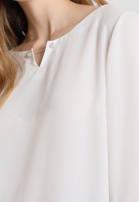 Valkoinen pusero, jossa on V-kaula-aukko ja kaksi nappikoristetta, pitkät hihat ja kevyt kangas, joka luo pehmeän ja sileän tekstuurin.