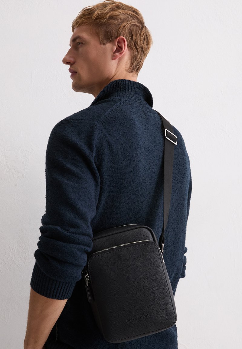 Homme en pull texturé bleu marine porte un sac bandoulière en cuir noir avec des fermetures éclair argentées sur fond blanc.
