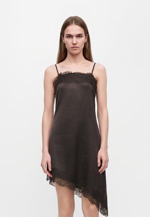 Jeune femme debout portant une robe nuisette noire bordée de dentelle superposée sur un jean flare bleu clair et des chaussures noires à bouts pointus.