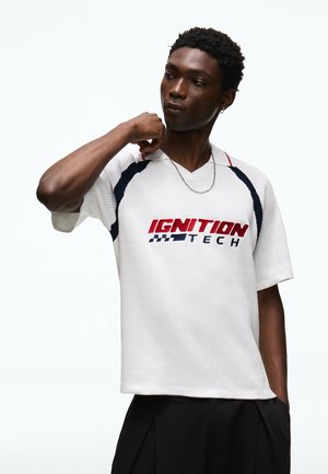 Jeune homme portant une chemise blanche avec le texte "IGNITION TECH", ajustant le col d'une main, sur un fond clair uni.