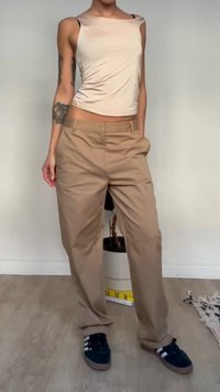 Person wearing loose beige trousers, cream sleeveless top, and dark blue sneakers standing indoors on wooden floor.