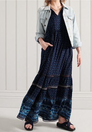 Kvinde iført en navyblå mønstret maxikjole, lys denimjakke med op-rullede ærmer, sorte sandaler og lag-på-lag guldhalskæder.