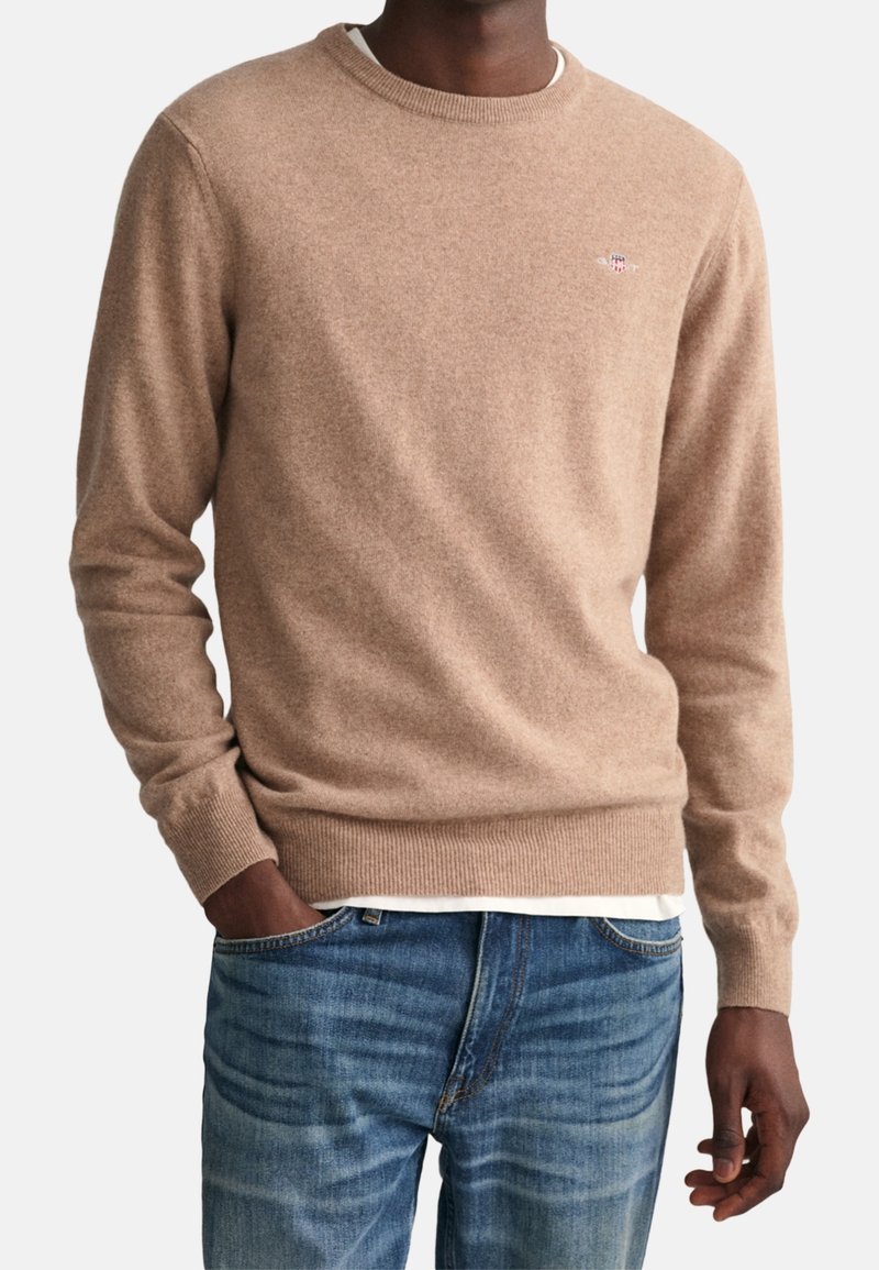 Homme portant un pull beige col rond par-dessus une chemise blanche avec un jean bleu, main gauche dans la poche, sur un fond uni.