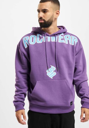 Lila Kapuzenpullover mit Kängurutasche, der "ROCKWEAR" in blauer Schrift auf der Brust zeigt und ein stilisiertes blaues Logodetail aufweist.