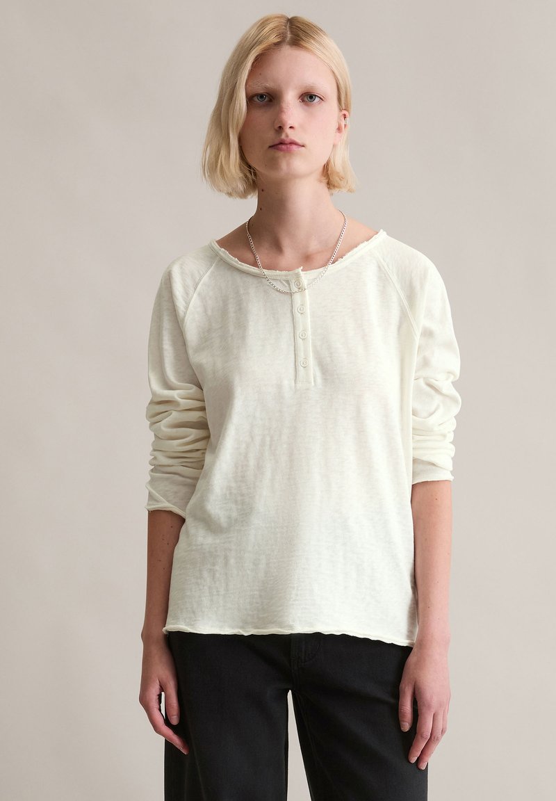 Langärmliges, off-white, leichtes Hemd mit einer Knopfleiste, rohem Saum und Raglanärmeln, kombiniert mit dunkler Hose. Glatte Textur.