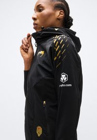 Μαύρο μπουφάν με κουκούλα από υφασμάτινη υφή, χρυσές λεπτομέρειες και τσέπες με φερμουάρ. Διαθέτει το λογότυπο UFC και τη σήμανση crypto.com στα μανίκια.