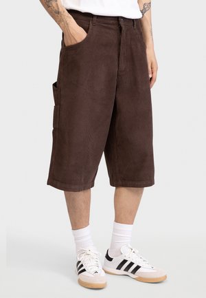 Άνδρας που φοράει καφέ βερμούδα κοτλέ μέχρι το γόνατο, λευκές κάλτσες μέχρι τον αστράγαλο και λευκά αθλητικά παπούτσια με μαύρες ρίγες, με τα χέρια εν μέρει στις τσέπες.