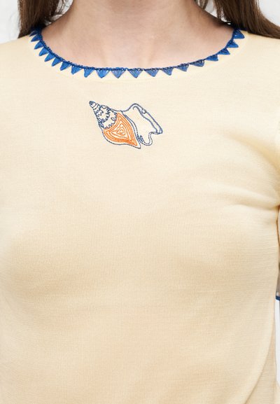 Μπεζ πουκάμισο με μπλε τρίγωνη διακόσμηση κατά μήκος της λαιμόκοψης και κεντημένο σχέδιο κοχυλιού σε μπλε και πορτοκαλί κοντά στον γιακά.