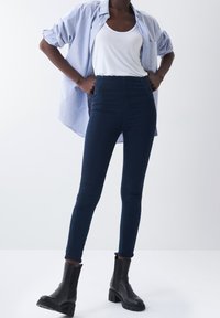 Höga mörk denim skinny jeans i kombination med en vit linne och en ljusblå skjorta med knappar, kompletterat med svarta ankelstövlar.