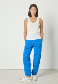 Junge Frau steht mit den Händen in den Taschen, trägt ein weißes Tanktop, leuchtend blaue Jogginghosen und weiße Leinensneaker.