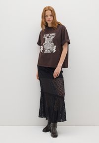 Camiseta gráfica marrón con diseño de gato y texto. Falda maxi de encaje negro transparente con detalles en capas. Modelo vistiendo botas negras hasta el tobillo.