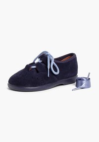 Scarpa in suede blu navy con dettagli perforati, suola in gomma piatta e lacci azzurri chiari. Un nastro per i lacci coordinato è posizionato accanto alla scarpa.