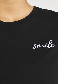 Černé bavlněné tričko s kulatým výstřihem a bílým vyšívaným nápisem "smile" na přední straně, detailní záběr na látku a detaily.