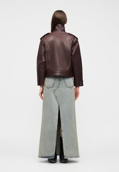 Άτομο με μακριά μαλλιά φοράει καφέ δερμάτινο μπουφάν και φούστα μακριά από ανοιχτό τζιν με ψηλή μέση και σκίσιμο στο πίσω μέρος, γυρισμένο με την πλάτη σε λευκό φόντο.