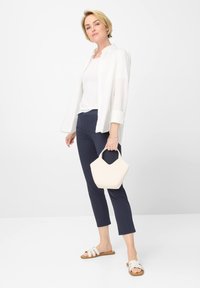 Camicia bianca a bottoni sopra una maglietta bianca, pantaloni blu navy e sandali bianchi. Porta una piccola borsa color crema con una forma unica.