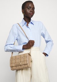 Borsa a tracolla intrecciata beige con un accento dorato e una tracolla removibile. Presenta una forma rettangolare con una texture liscia.