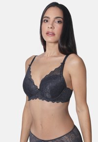 Schwarzer Spitzen-BH mit gepolsterten Cups, floralem Muster, gezacktem Rand, verstellbaren Trägern und einer dekorativen Schleife in der Mitte. Glatter Stoff auf der Rückseite.