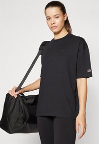 Svart bomulls t-skjorte med korte ermer og rund hals, med en liten logo på brystet og brukt sammen med en svart treningsbag.