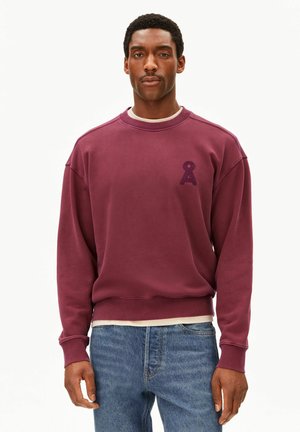 Homme portant un sweat-shirt bordeaux à col rond avec une petite lettre A texturée sur la poitrine et un jean bleu, debout devant un fond uni.