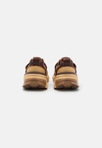 Bruna och beige idrottsskor med en strukturerad gummisula, vadderad häl och ett justerbart remdesign. Baksidan visar detaljer.