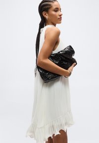 Kobieta z długimi warkoczami, ubrana w białą, bezrękawną sukienkę, trzyma dużą, czarną, teksturowaną kopertówkę na jednolitym tle.