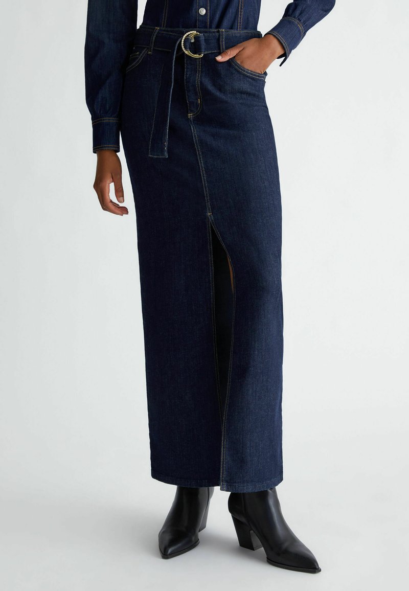 Gonna maxi in denim blu scuro con spacco frontale, vita con cintura, accessori dorati e cuciture marrone chiaro. Abbinata a stivali neri con tacco.