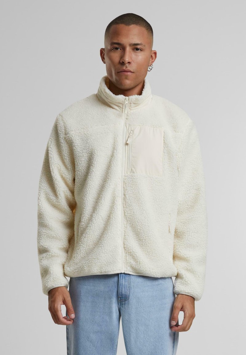 Giacca in pile crema con colletto alto, chiusura con zip e tasca frontale. Tessuto morbido, vestibilità rilassata, abbinata a jeans di denim azzurro.