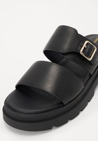 Sandale slide noire avec deux larges brides, dont une avec une boucle dorée, et une semelle épaisse texturée.