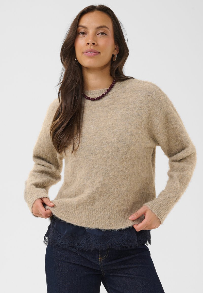 Femme aux longs cheveux bruns porte un pull beige duveteux, un jean foncé, un top en dentelle bleu marine en dessous et un collier de perles, les mains sur l’ourlet du pull.