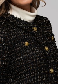 Frau trägt schwarze, strukturierte Jacke mit Goldfäden und dekorativen goldenen Knöpfen über cremefarbenem Rollkragenpullover.