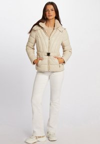 Giacca beige trapuntata con cappuccio, caratterizzata da un colletto morbido, vita con cintura e chiusura con zip. Abbinata a pantaloni bianchi e scarpe chiare.