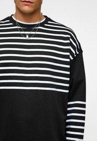 Zwarte langearmige shirt met horizontale witte strepen, met een ronde halslijn en een ontspannen pasvorm. Inclusief metallic gelaagde halskettingen.