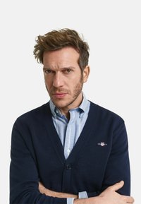 Marinefarbener Cardigan aus strukturiertem Strick mit V-Ausschnitt und Knopfschluss, getragen über einem hellblau gestreiften Hemd. Mit Logodetail auf der Brust.