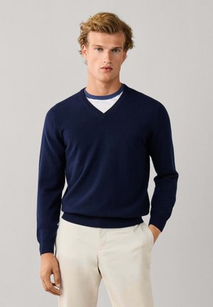 Joven con cabello rubio que lleva un jersey azul marino con cuello en V sobre una camisa blanca y pantalones beige, de pie con una mano en el bolsillo.
