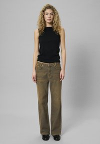 Czarny bezrękawnik z wysokim dekoltem, dopasowany do wyblakłych brązowych spodni z szerokimi nogawkami. Modelka stoi na szarym tle, w czarnych butach.