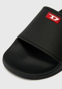 Black slide sandal with a textured rubber upper and a flat sole. Features a red logo accent. Smooth, matte finish.
