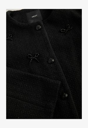 Schwarze, strukturierte Strickjacke mit rundem Halsausschnitt, gemustertem Stoff, schwarzen Knöpfen und dekorativen Perlenakzenten in Form von Schleifen.