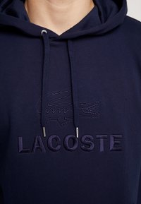 Námořnický modrý mikina s kapucí na šňůrku, s vyšitým logo krokodýla a textem "LACOSTE" ve stejné barvě. Texturovaná látka.