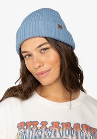 Berretto a coste blu chiaro con una texture morbida e una piccola etichetta logata marrone. Indossato con una t-shirt bianca con grafica e testo colorato.