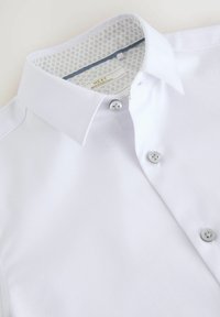 Camicia bianca con colletto classico, caratterizzata da bottoni argentati e una fodera interna a pattern. Realizzata in tessuto liscio e tessuto a mano.