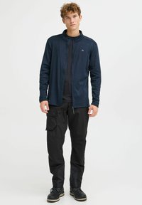 Marineblauer Reißverschlussjacke aus strukturiertem Stoff mit hohem Kragen, kombiniert mit schwarzen Cargo-Hosen mit Taschen. Schwarze Schuhe runden den Look ab.