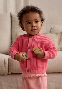 Cardigan rosa lavorato a maglia con trama a coste e bottoni in legno, abbinato a pantaloni rosa a quadretti. Bambino che tiene in mano dei blocchi di legno, seduto su un divano.